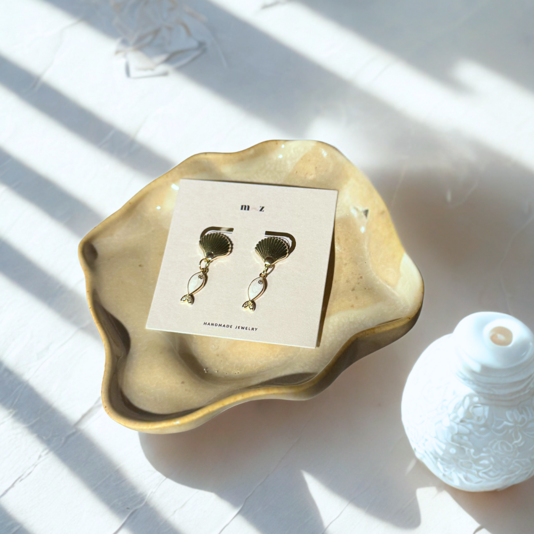 Gold earrings on a card with a decorative dish and shell in the background